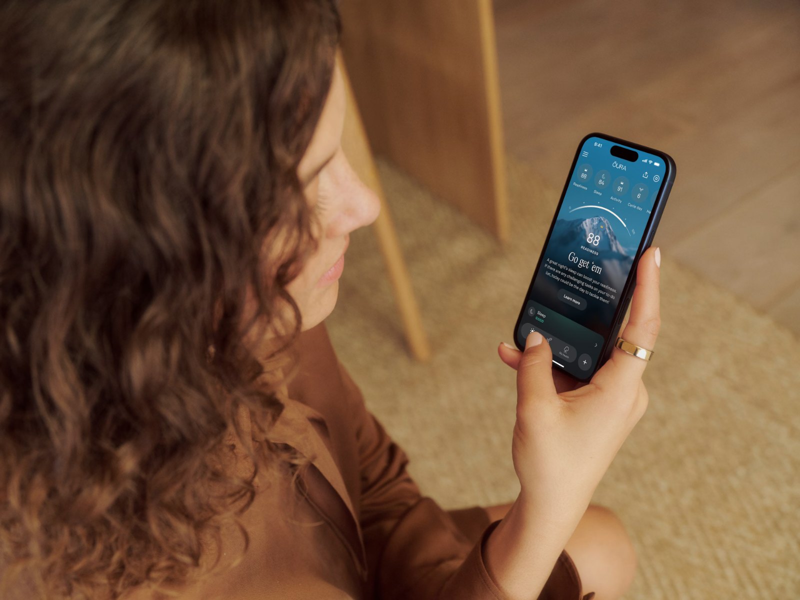 Une femme aux cheveux bouclés, la KEORA Ring 5 au doigt, consulte l’application KEORA sur un smartphone.